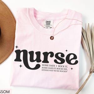 Pode incluir: Camiseta rosa claro com a palavra "nurse" em preto, com uma fonte retr&ocirc;. A camiseta tamb&eacute;m apresenta o texto "SOME DAYS I ROCK IT, SOME DAYS IT ROCKS ME, EITHER WAY WE'RE ROCKIN'". Um colar dourado e um chap&eacute;u marrom tamb&eacute;m s&atilde;o vis&iacute;veis.