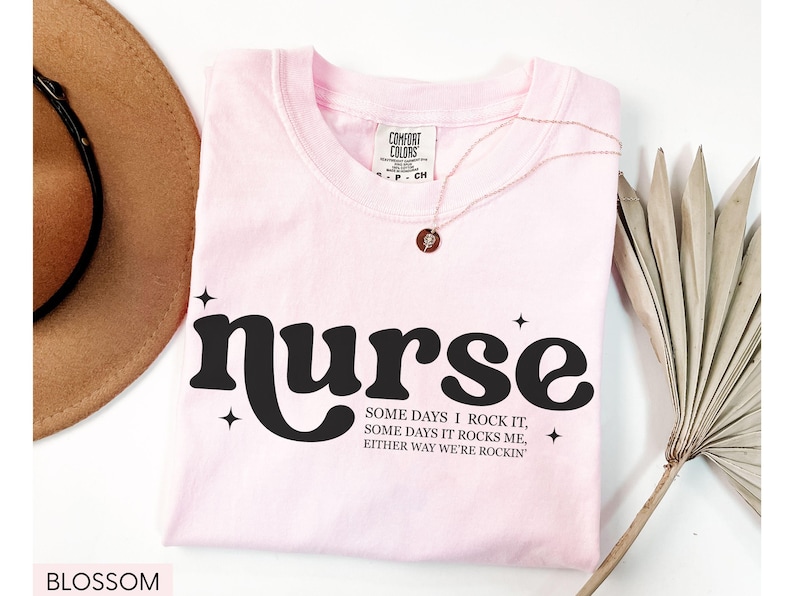 Pode incluir: Camiseta rosa claro com a palavra "nurse" em preto, com uma fonte retr&ocirc;. A camiseta tamb&eacute;m apresenta o texto "SOME DAYS I ROCK IT, SOME DAYS IT ROCKS ME, EITHER WAY WE'RE ROCKIN'". Um colar dourado e um chap&eacute;u marrom tamb&eacute;m s&atilde;o vis&iacute;veis.