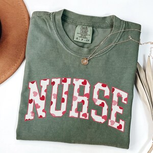 Pode incluir: Uma camiseta verde com a palavra "NURSE" impressa em letras brancas com um padr&atilde;o de cora&ccedil;&otilde;es vermelhos. A camiseta tem um visual desgastado e &eacute; feita de um tecido macio e confort&aacute;vel.