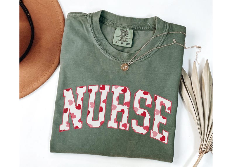 Pode incluir: Uma camiseta verde com a palavra "NURSE" impressa em letras brancas com um padr&atilde;o de cora&ccedil;&otilde;es vermelhos. A camiseta tem um visual desgastado e &eacute; feita de um tecido macio e confort&aacute;vel.