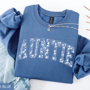 Peut inclure: Sweat-shirt bleu indigo avec le mot "AUNTIE" en imprim&eacute; floral blanc. Un collier en argent avec un pendentif est drap&eacute; sur le sweat-shirt. Le sweat-shirt est pli&eacute; sur une paire de jeans bleus.