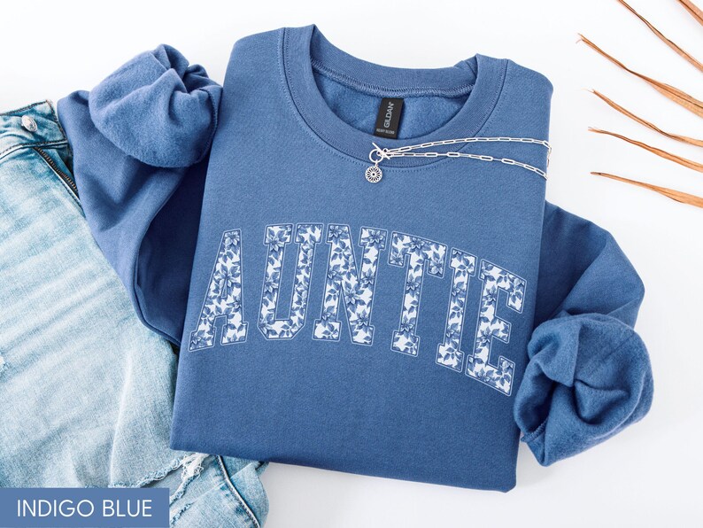 Peut inclure: Sweat-shirt bleu indigo avec le mot "AUNTIE" en imprim&eacute; floral blanc. Un collier en argent avec un pendentif est drap&eacute; sur le sweat-shirt. Le sweat-shirt est pli&eacute; sur une paire de jeans bleus.
