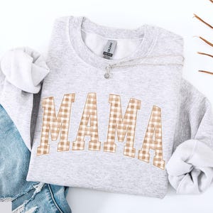 May include: A light gray sweatshirt with the word "MAMA" in large, arched letters. The letters have a tan and white gingham pattern. A silver necklace with a small pendant rests on the sweatshirt. The sleeves are cuffed, and a pair of blue jeans are visible.