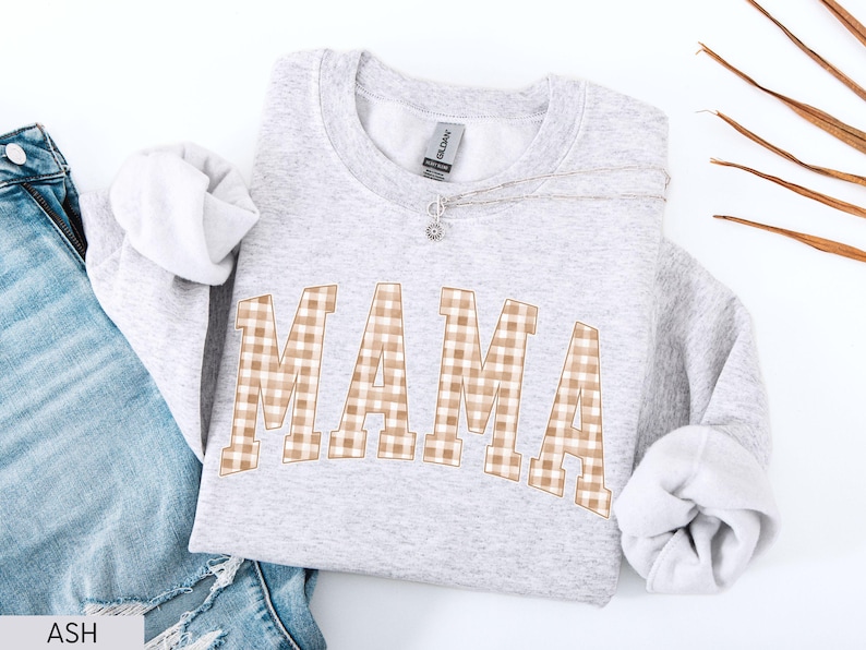 May include: A light gray sweatshirt with the word "MAMA" in large, arched letters. The letters have a tan and white gingham pattern. A silver necklace with a small pendant rests on the sweatshirt. The sleeves are cuffed, and a pair of blue jeans are visible.