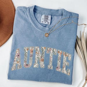 May include: A light blue t-shirt with the word "AUNTIE" in white letters outlined with a floral pattern. The shirt is made of a soft, comfortable material.