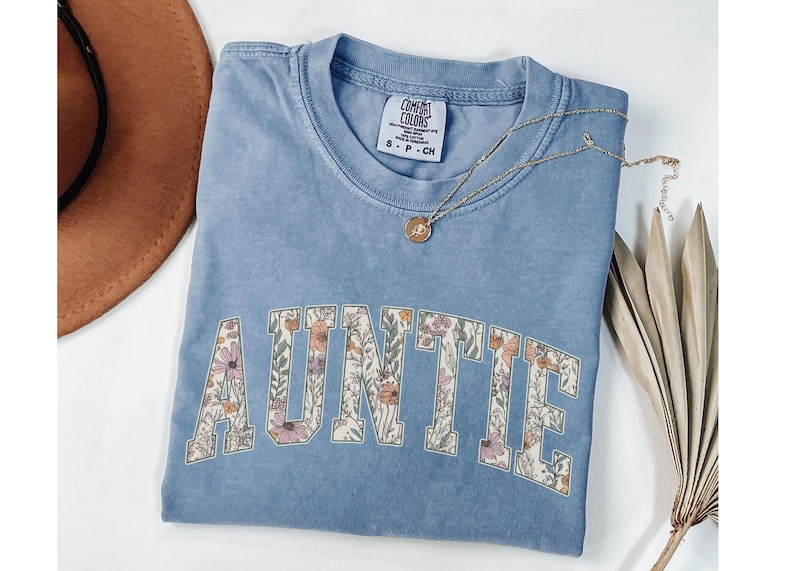 May include: A light blue t-shirt with the word "AUNTIE" in white letters outlined with a floral pattern. The shirt is made of a soft, comfortable material.