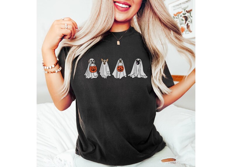 Puede incluir: Camiseta negra con un dise&ntilde;o de Halloween. La camiseta muestra cuatro perros disfrazados de fantasmas, cada uno sosteniendo una calabaza. El dise&ntilde;o es en blanco, naranja y negro. La camiseta es de estilo casual.
