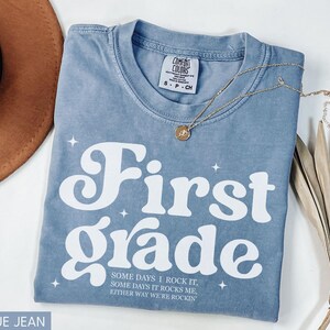Puede incluir: Camiseta azul vaquero con las palabras "First Grade" en blanco, junto con el texto "SOME DAYS I ROCK IT, SOME DAYS IT ROCKS ME, EITHER WAY WE'RE ROCKIN". Tambi&eacute;n se ven un collar dorado y un sombrero marr&oacute;n.