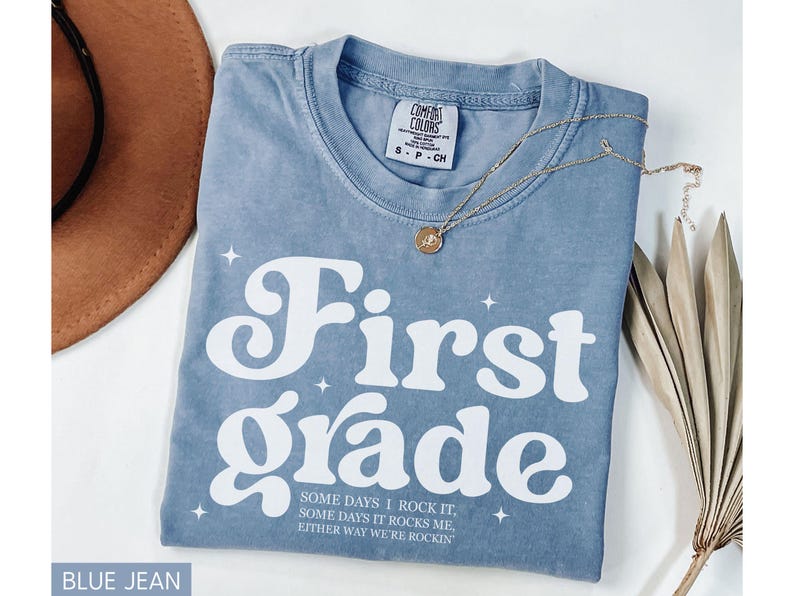 Puede incluir: Camiseta azul vaquero con las palabras "First Grade" en blanco, junto con el texto "SOME DAYS I ROCK IT, SOME DAYS IT ROCKS ME, EITHER WAY WE'RE ROCKIN". Tambi&eacute;n se ven un collar dorado y un sombrero marr&oacute;n.