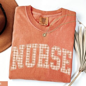 May include: A coral-colored t-shirt with the word "NURSE" in a plaid pattern across the front. A gold necklace with a pendant is draped on the shirt. A brown hat and a dried palm leaf are in the background.
