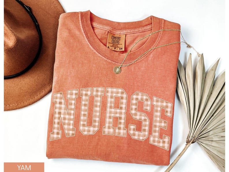 May include: A coral-colored t-shirt with the word "NURSE" in a plaid pattern across the front. A gold necklace with a pendant is draped on the shirt. A brown hat and a dried palm leaf are in the background.