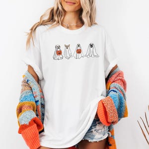 Puede incluir: Camiseta blanca con un dise&ntilde;o de Halloween que presenta cuatro perros disfrazados de fantasmas. Dos perros sostienen calabazas. La camiseta es de estilo casual y unisex. La modelo lleva un su&eacute;ter colorido.