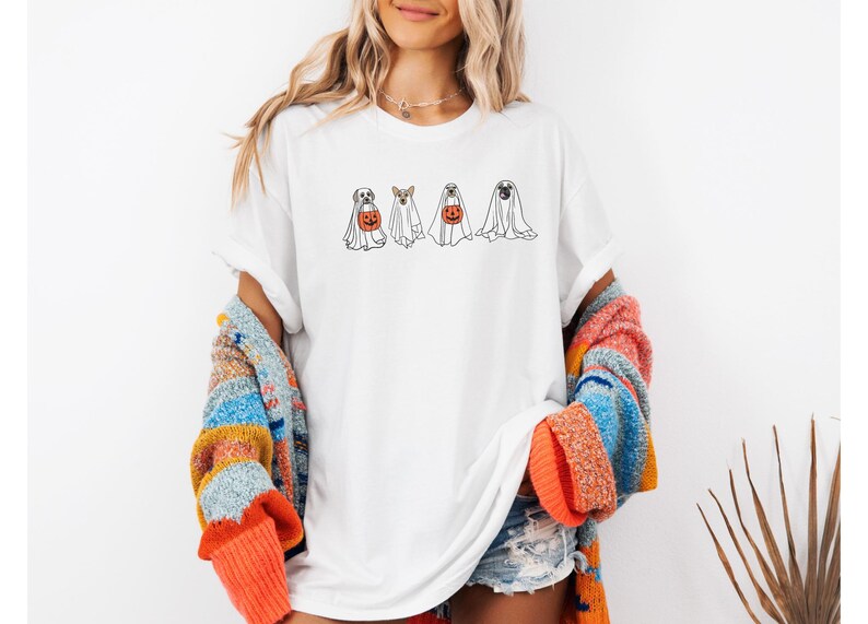 Puede incluir: Camiseta blanca con un dise&ntilde;o de Halloween que presenta cuatro perros disfrazados de fantasmas. Dos perros sostienen calabazas. La camiseta es de estilo casual y unisex. La modelo lleva un su&eacute;ter colorido.