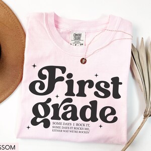Puede incluir: Una camiseta rosa claro con las palabras "First Grade" en una fuente audaz y retro. La camiseta tambi&eacute;n presenta el texto "SOME DAYS I ROCK IT, SOME DAYS IT ROCKS ME, EITHER WAY WE'RE ROCKIN'". Tambi&eacute;n se ven un collar dorado y un sombrero marr&oacute;n.