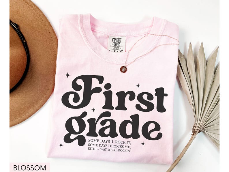 Puede incluir: Una camiseta rosa claro con las palabras "First Grade" en una fuente audaz y retro. La camiseta tambi&eacute;n presenta el texto "SOME DAYS I ROCK IT, SOME DAYS IT ROCKS ME, EITHER WAY WE'RE ROCKIN'". Tambi&eacute;n se ven un collar dorado y un sombrero marr&oacute;n.