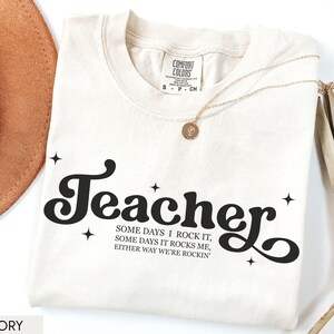 May include: Ivory-colored t-shirt with the word "Teacher" in a black, retro font. The shirt also features the text "SOME DAYS I ROCK IT, SOME DAYS IT ROCKS ME, EITHER WAY WE'RE ROCKIN". A gold necklace and a brown hat are also visible.