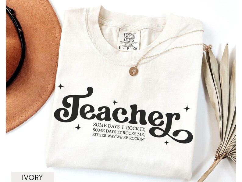 May include: Ivory-colored t-shirt with the word "Teacher" in a black, retro font. The shirt also features the text "SOME DAYS I ROCK IT, SOME DAYS IT ROCKS ME, EITHER WAY WE'RE ROCKIN". A gold necklace and a brown hat are also visible.