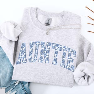 Peut inclure: Un sweat-shirt gris clair avec le mot "AUNTIE" en grandes lettres florales bleues sur la poitrine. Le sweat-shirt est pli&eacute; avec les manches rentr&eacute;es et un collier argent&eacute; est drap&eacute; sur le devant. Un jean bleu est visible en arri&egrave;re-plan.