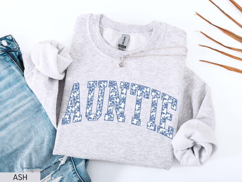 Peut inclure: Un sweat-shirt gris clair avec le mot "AUNTIE" en grandes lettres florales bleues sur la poitrine. Le sweat-shirt est pli&eacute; avec les manches rentr&eacute;es et un collier argent&eacute; est drap&eacute; sur le devant. Un jean bleu est visible en arri&egrave;re-plan.