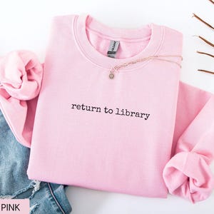 May include: A light pink sweatshirt with the text "return to library" in black. The sweatshirt is folded with the sleeves rolled up. A gold necklace with a small pendant is on top of the sweatshirt. Blue jeans are visible in the background.