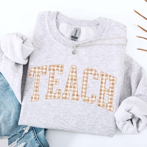 May include: A light gray sweatshirt with the word "TEACH" in a brown and white plaid pattern across the front. The sweatshirt is folded with the sleeves showing. A silver necklace with a small pendant is on the sweatshirt. Blue jeans are visible in the background.