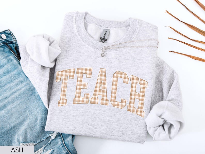 May include: A light gray sweatshirt with the word "TEACH" in a brown and white plaid pattern across the front. The sweatshirt is folded with the sleeves showing. A silver necklace with a small pendant is on the sweatshirt. Blue jeans are visible in the background.