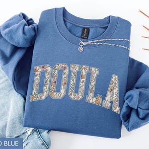 May include: Indigo blue sweatshirt with the word "DOULA" in a floral design. A silver necklace with a small pendant rests on the sweatshirt. The sweatshirt is folded on top of a pair of blue jeans.