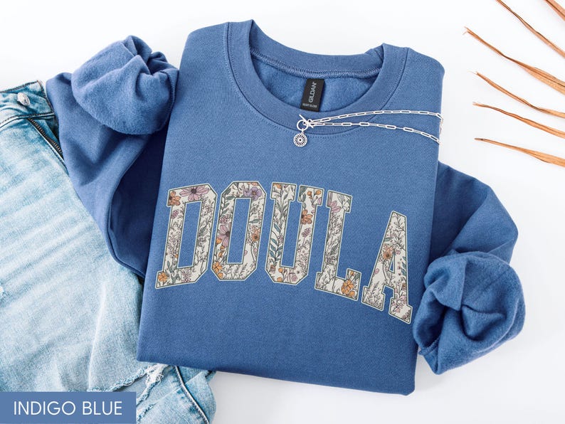 May include: Indigo blue sweatshirt with the word "DOULA" in a floral design. A silver necklace with a small pendant rests on the sweatshirt. The sweatshirt is folded on top of a pair of blue jeans.
