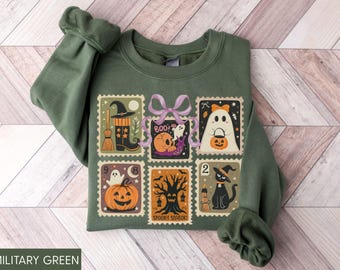 May include: Military green sweatshirt featuring a Halloween-themed design. The design includes six postage stamp-style illustrations with a witch's hat, ghost, pumpkin, and black cat. The text "BOO!" and "Spooky Season" are also included.