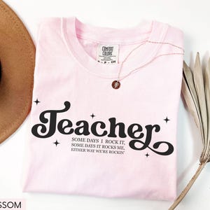 May include: A light pink t-shirt with the word "Teacher" in a black, retro font. The shirt also features the text "SOME DAYS I ROCK IT, SOME DAYS IT ROCKS ME, EITHER WAY WE'RE ROCKIN". A gold necklace and a brown hat are also visible.