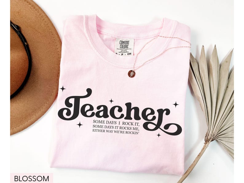 May include: A light pink t-shirt with the word "Teacher" in a black, retro font. The shirt also features the text "SOME DAYS I ROCK IT, SOME DAYS IT ROCKS ME, EITHER WAY WE'RE ROCKIN". A gold necklace and a brown hat are also visible.