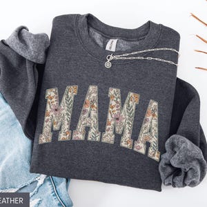 May include: Dark heather gray sweatshirt with the word "MAMA" in a floral design across the front. A silver necklace is draped across the sweatshirt. The sweatshirt is folded next to a pair of blue jeans.