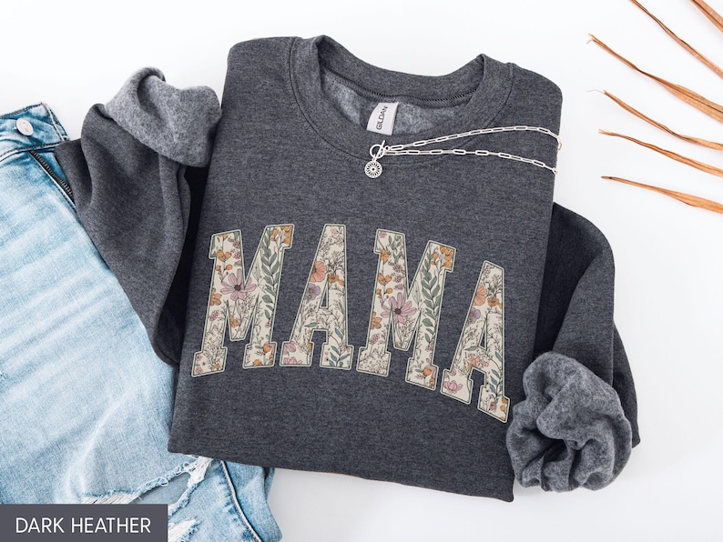 May include: Dark heather gray sweatshirt with the word "MAMA" in a floral design across the front. A silver necklace is draped across the sweatshirt. The sweatshirt is folded next to a pair of blue jeans.