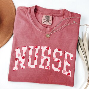 Pode incluir: Uma camiseta vermelha com a palavra "NURSE" impressa em letras brancas com um padr&atilde;o de cora&ccedil;&otilde;es rosa e vermelhos. A camiseta &eacute; feita de um material macio e confort&aacute;vel.