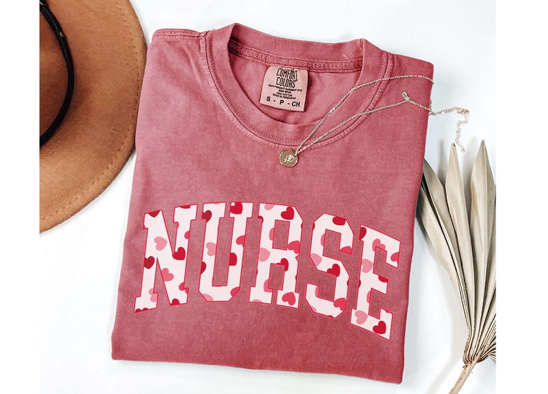 Pode incluir: Uma camiseta vermelha com a palavra "NURSE" impressa em letras brancas com um padr&atilde;o de cora&ccedil;&otilde;es rosa e vermelhos. A camiseta &eacute; feita de um material macio e confort&aacute;vel.