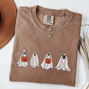 Puede incluir: Camiseta Comfort Colors marr&oacute;n con un dise&ntilde;o de cuatro perros disfrazados de fantasmas, cada uno sosteniendo una calabaza. La camiseta tiene cuello redondo y mangas cortas. Un collar plateado con un peque&ntilde;o colgante est&aacute; sobre la camiseta. Un sombrero marr&oacute;n est&aacute; en el fondo.