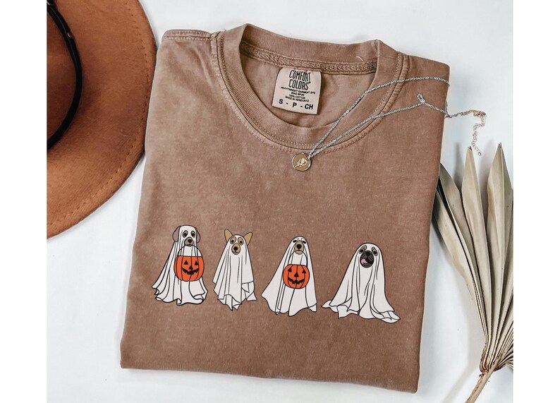 Puede incluir: Camiseta Comfort Colors marr&oacute;n con un dise&ntilde;o de cuatro perros disfrazados de fantasmas, cada uno sosteniendo una calabaza. La camiseta tiene cuello redondo y mangas cortas. Un collar plateado con un peque&ntilde;o colgante est&aacute; sobre la camiseta. Un sombrero marr&oacute;n est&aacute; en el fondo.