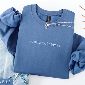 May include: Indigo blue sweatshirt with the text "return to library" in white. A silver necklace with a small pendant is draped across the top. The sweatshirt is folded on top of a pair of blue jeans.