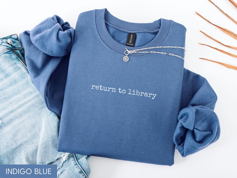 May include: Indigo blue sweatshirt with the text "return to library" in white. A silver necklace with a small pendant is draped across the top. The sweatshirt is folded on top of a pair of blue jeans.