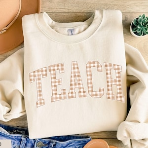 May include: Cream-colored sweatshirt with the word "TEACH" in a brown and white gingham pattern across the front. The sweatshirt is paired with blue jeans and a tan hat. A small succulent plant in a white pot is visible.