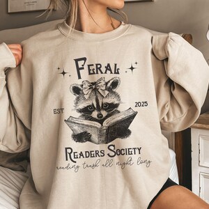 May include: A beige sweatshirt with a black graphic of a raccoon reading a book. The graphic includes the text "Feral Readers Society" and "reading trash all night long".