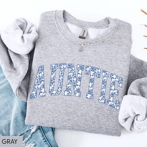 Peut inclure: Un sweat-shirt gris clair avec le mot "AUNTIE" imprim&eacute; en bleu floral. Le sweat-shirt est pli&eacute; &agrave; c&ocirc;t&eacute; d'un jean bleu. Le texte "SPORT GRAY" est visible en bas &agrave; gauche.