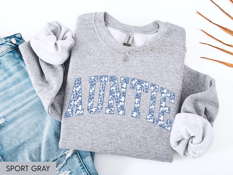 Peut inclure: Un sweat-shirt gris clair avec le mot "AUNTIE" imprim&eacute; en bleu floral. Le sweat-shirt est pli&eacute; &agrave; c&ocirc;t&eacute; d'un jean bleu. Le texte "SPORT GRAY" est visible en bas &agrave; gauche.