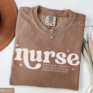 Pode incluir: Camiseta cor espresso com a palavra "nurse" em uma fonte retr&ocirc;, acompanhada do texto "Some days I rock it, some days it rocks me, either way we're rockin'". Um colar de prata com um pingente est&aacute; na camiseta.