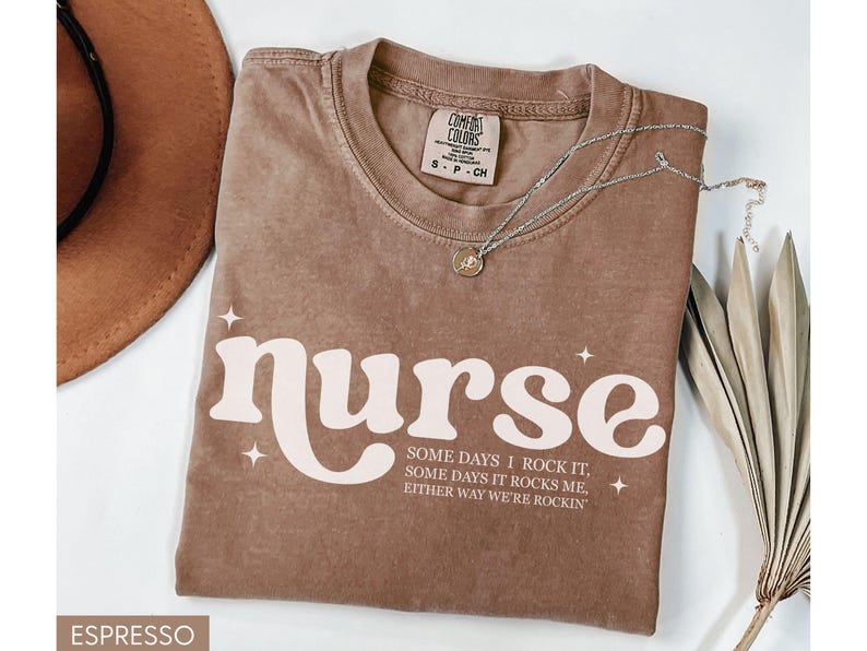 Pode incluir: Camiseta cor espresso com a palavra "nurse" em uma fonte retr&ocirc;, acompanhada do texto "Some days I rock it, some days it rocks me, either way we're rockin'". Um colar de prata com um pingente est&aacute; na camiseta.