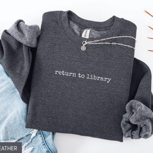 May include: Dark heather gray sweatshirt with the text "return to library" in white. A silver necklace with a circular pendant is draped across the sweatshirt. The sweatshirt is paired with blue jeans.