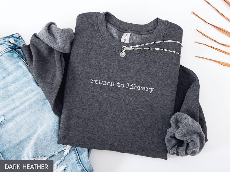 May include: Dark heather gray sweatshirt with the text "return to library" in white. A silver necklace with a circular pendant is draped across the sweatshirt. The sweatshirt is paired with blue jeans.