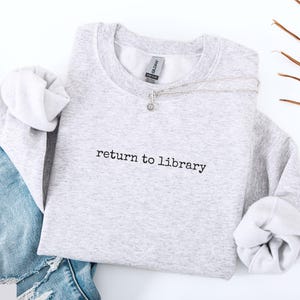 May include: A light gray sweatshirt with the text "return to library" in black. The sweatshirt is paired with blue jeans and a silver necklace. The sleeves are cuffed, and the overall aesthetic is casual and comfortable.
