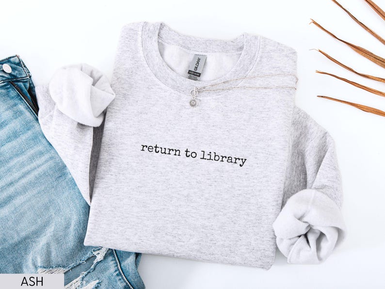 May include: A light gray sweatshirt with the text "return to library" in black. The sweatshirt is paired with blue jeans and a silver necklace. The sleeves are cuffed, and the overall aesthetic is casual and comfortable.