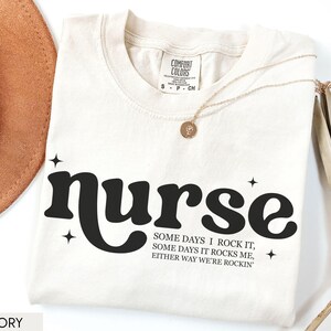 Pode incluir: Camiseta cor marfim com a palavra "nurse" em negrito preto, acompanhada do texto "Some days I rock it, some days it rocks me, either way we're rockin'". Um colar dourado e um chap&eacute;u marrom tamb&eacute;m s&atilde;o vis&iacute;veis.